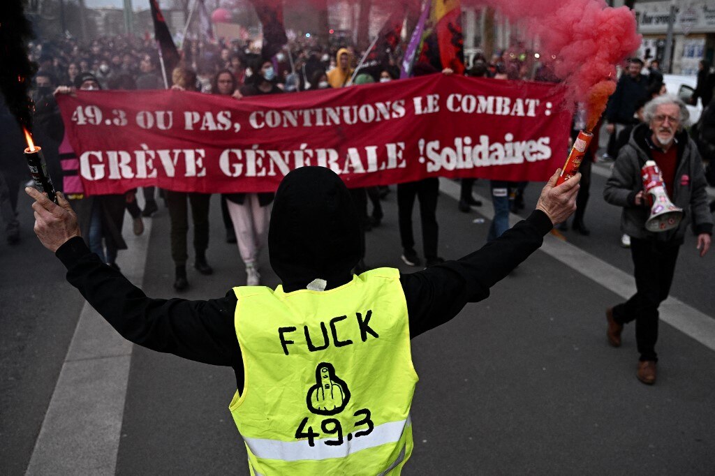 प्रदर्शनकारी सॉलिडेयर्स ट्रेड यूनियन रीडिंग का बैनर पकड़े हुए हैं "49.3 या नहीं, चलो लड़ते रहें, सामान्य हड़ताल" पीली बनियान पहने एक प्रदर्शनकारी के सामने पढ़ना "भाड़ में जाओ 49.3" 16 मार्च, 2023 को नैनटेस, पश्चिमी फ़्रांस में, संविधान के अनुच्छेद 49.3 का उपयोग करते हुए, फ्रांसीसी सरकार द्वारा संसद के माध्यम से एक वोट के बिना पेंशन सुधार को आगे बढ़ाने के बाद एक प्रदर्शन के दौरान। फ्रांसीसी राष्ट्रपति ने 16 मार्च को संसद के माध्यम से एक विवादास्पद पेंशन सुधार को आगे बढ़ाया। वोट के बिना, शायद ही कभी इस्तेमाल की जाने वाली संवैधानिक शक्ति को तैनात करना जो विरोध प्रदर्शनों को भड़काने का जोखिम उठाता है।  यह कदम एक प्रवेश था कि उनकी सरकार के पास सेवानिवृत्ति की आयु 62 से बढ़ाकर 64 करने के लिए कानून पारित करने के लिए नेशनल असेंबली में बहुमत की कमी थी। (LOIC VENANCE / AFP)